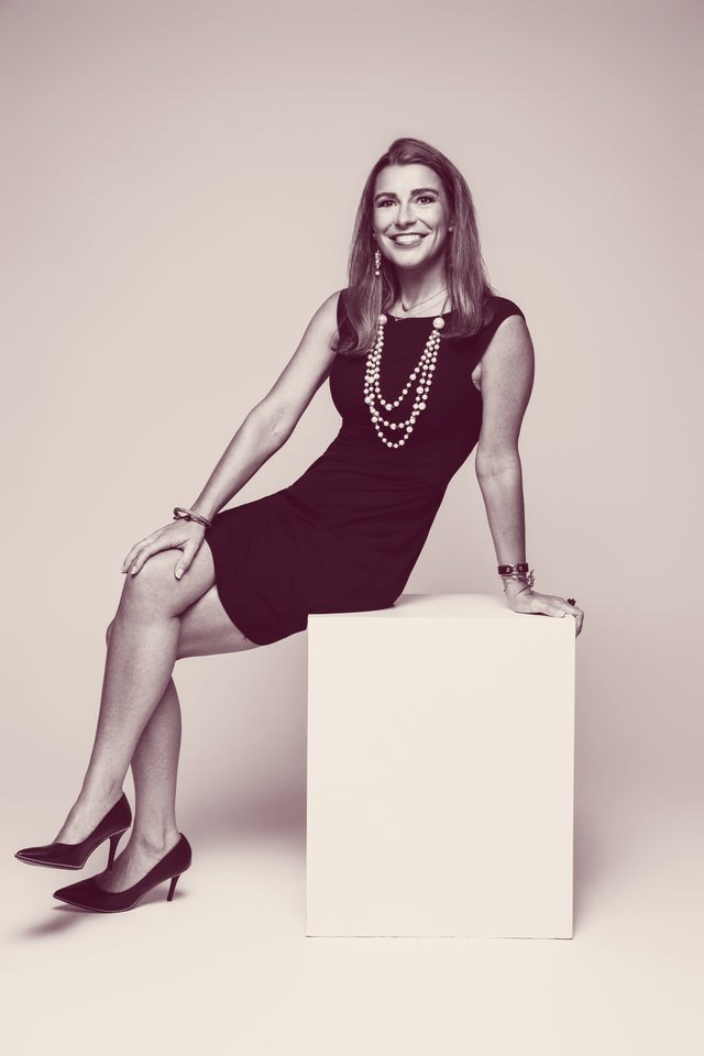 Sarah N. Dimmich in a black dress and pearls, seated on a white cube